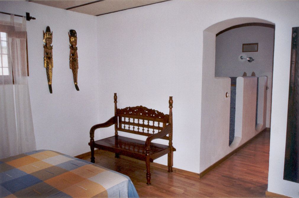 A bedroom corner with a carved wooden bench, decorative masks, and an archway leading to the ensuite bathroom