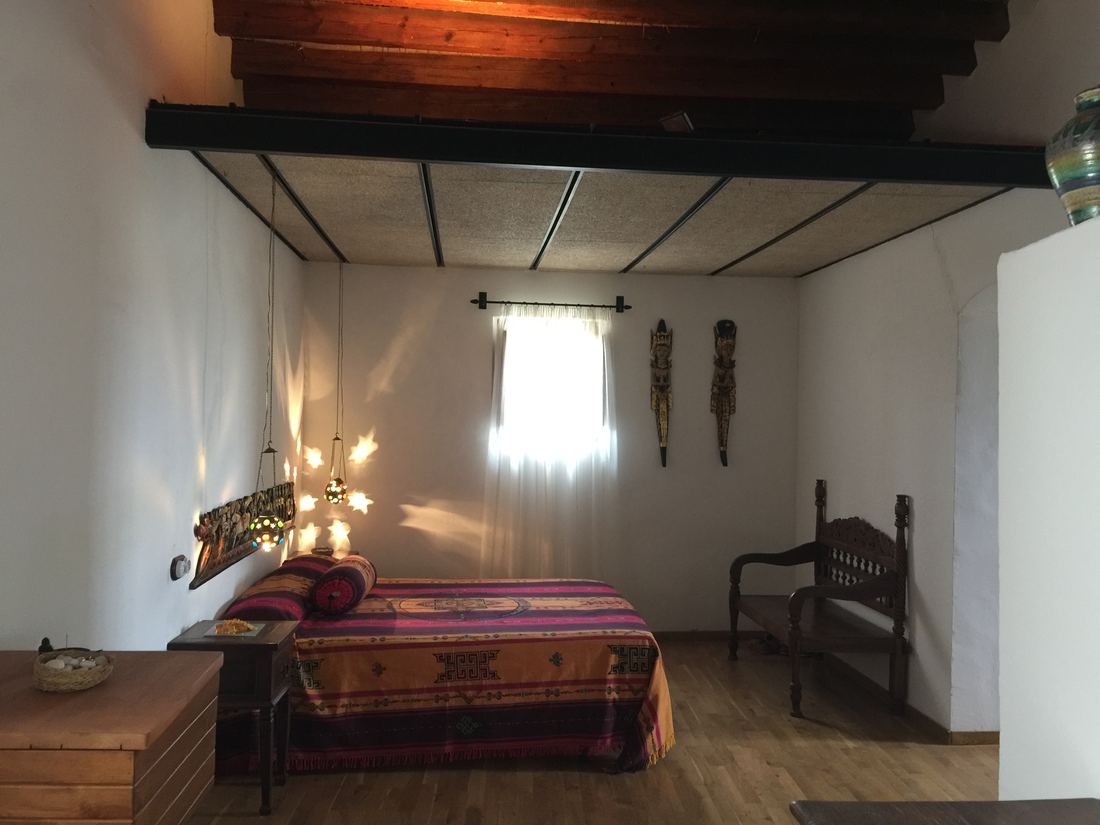 A bedroom with a patterned bedspread, carved wall hanging, hanging lanterns and decorative wooden masks